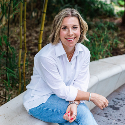 picture shows nikki butler, a white woman with a blonde bob sitting on a white concrete ledge wearing jeans and a white shirt and trainers