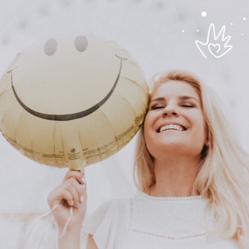 Woman in a white top with blonde hair holding a yellow balloon with a smile on it.